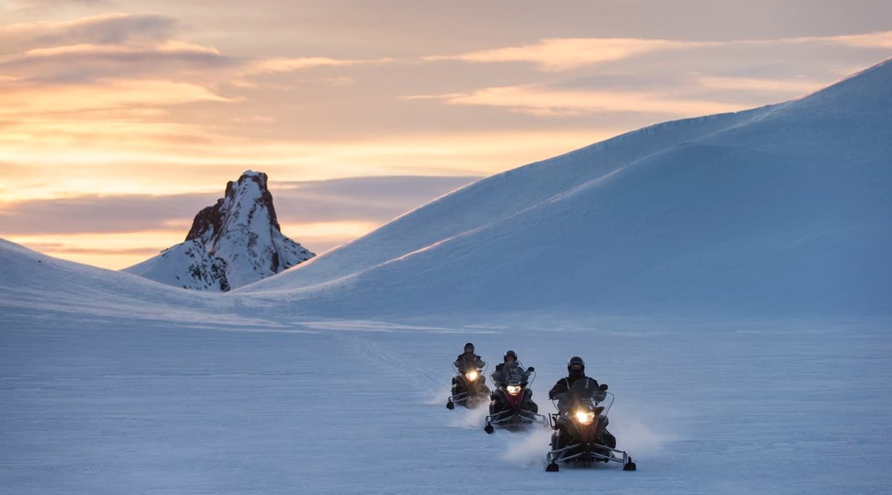 Drei Schneemobile überqueren einen isländischen Gletscher bei Sonnenuntergang.