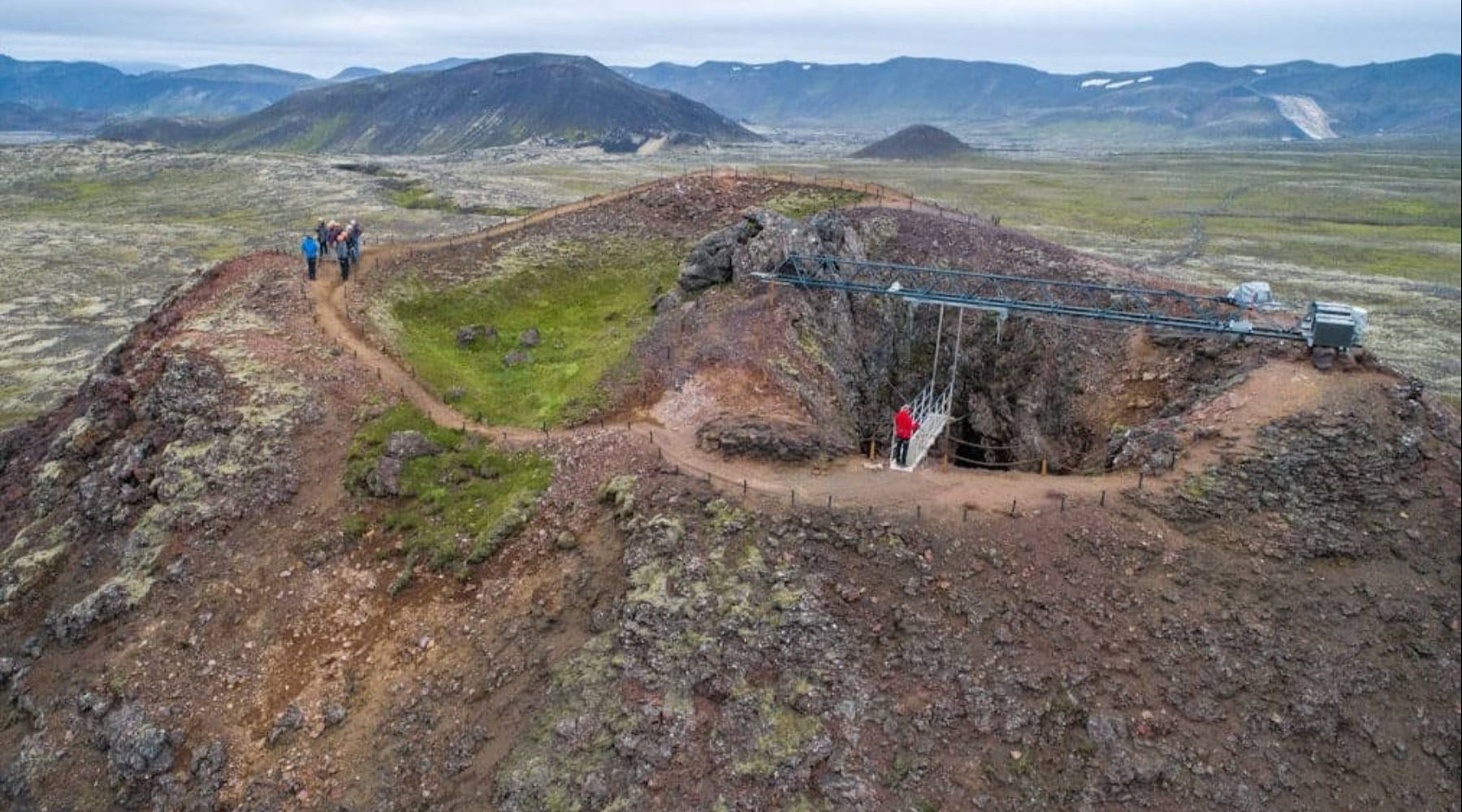 Hikers walking around a volcanic crater with a bridge over the entrance to the chamber