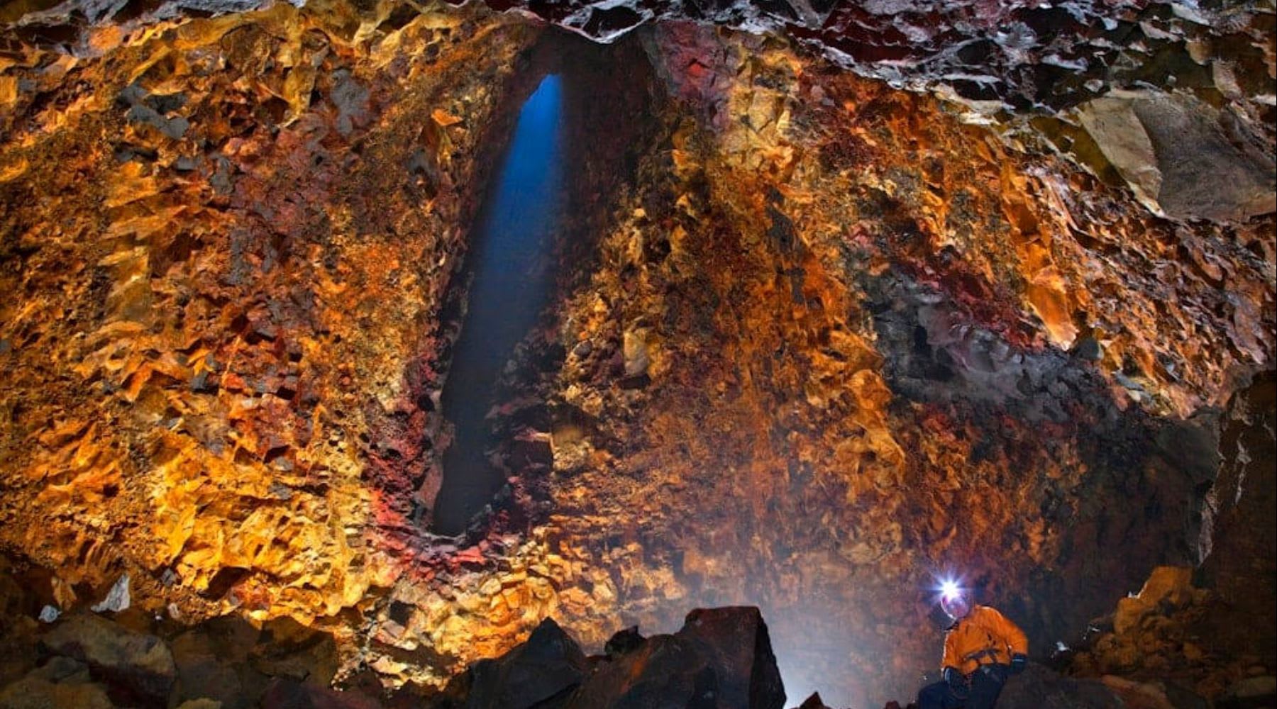 Illuminated volcanic chamber with colorful rock walls and a visitor wearing a headlamp