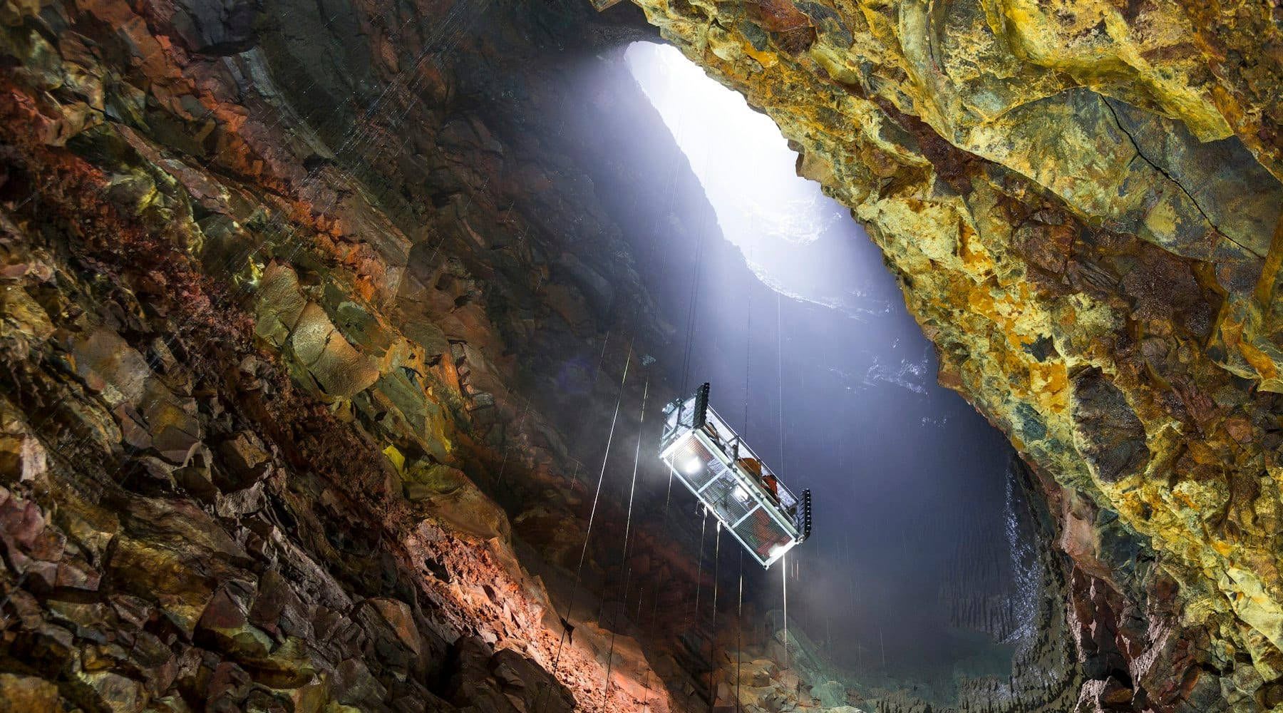 Metal lift descending into a deep volcanic chamber with light shining through the crater opening