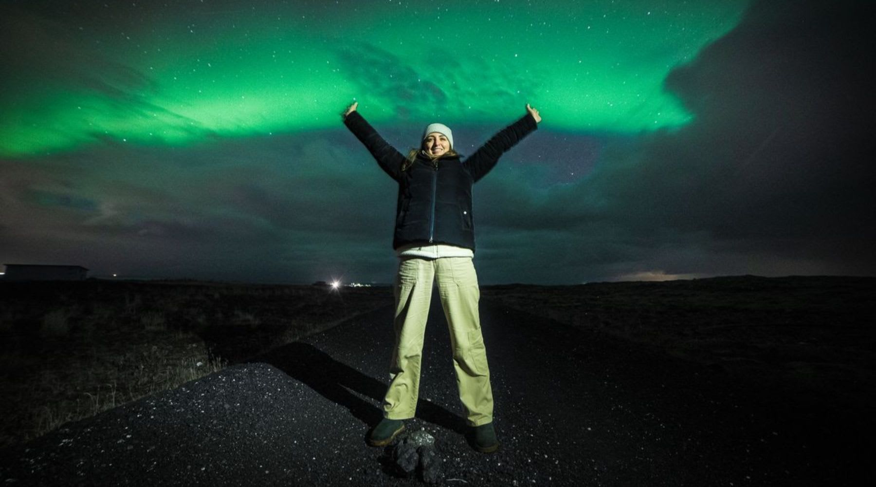 Traveler posing with arms raised beneath green northern lights in Iceland