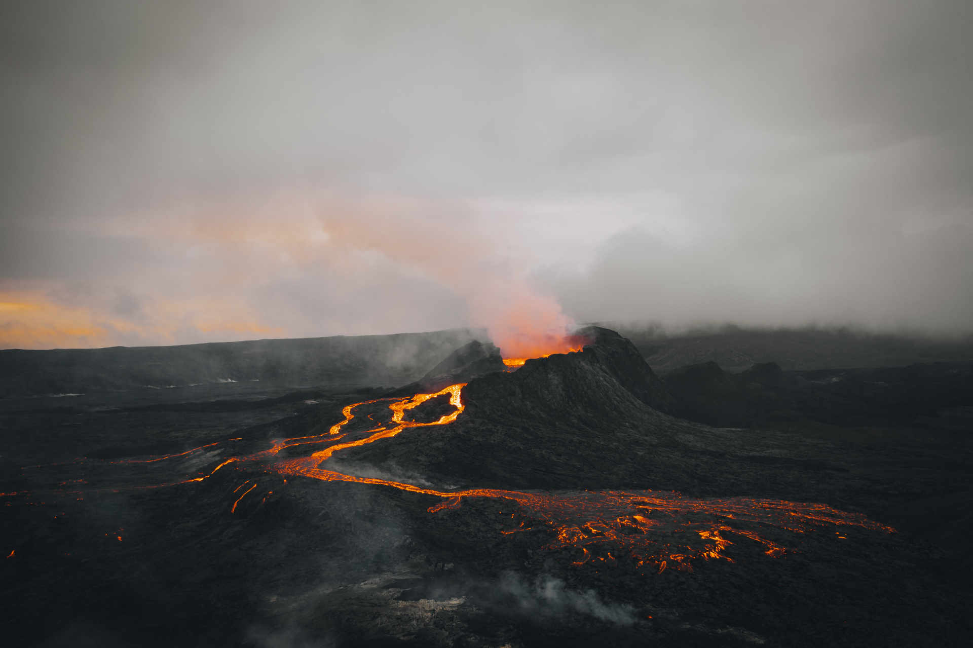 Aerial view of an erupting volcano at dusk with glowing lava flowing across dark terrain