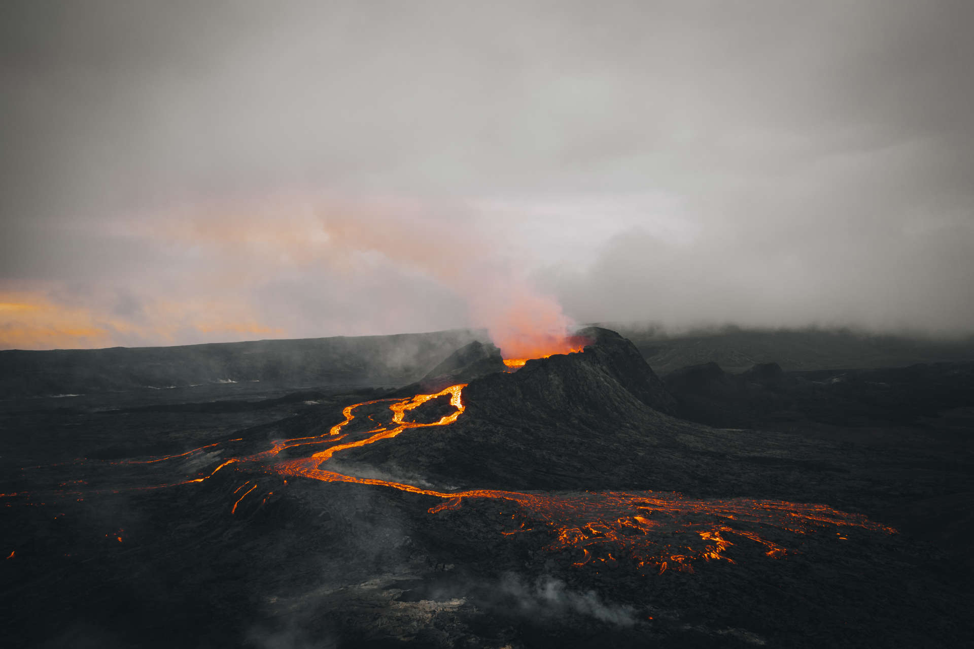 Aerial view of an erupting volcano at dusk with glowing lava flowing across dark terrain