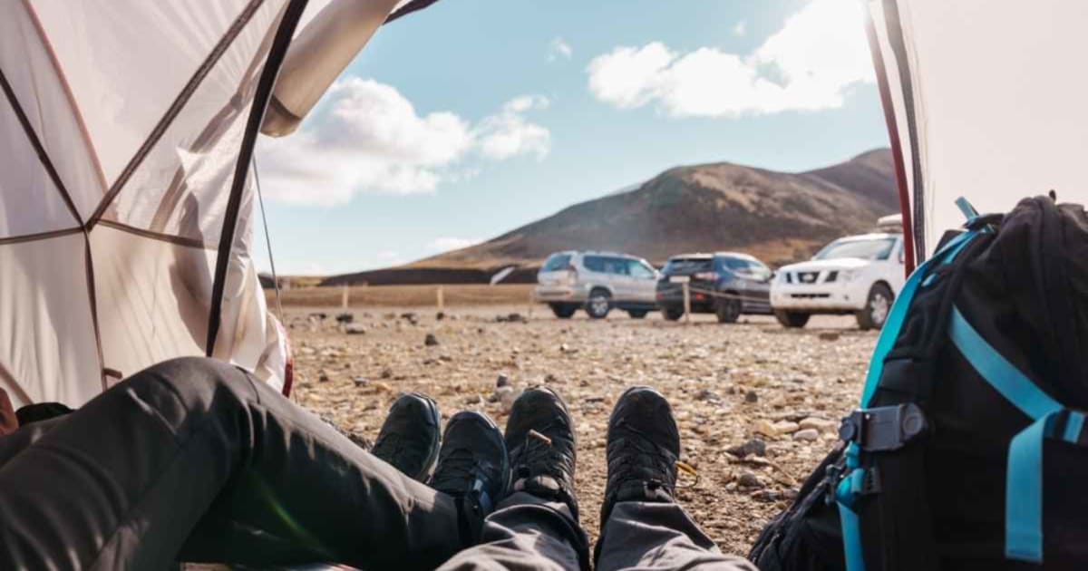 Traveler relaxing inside a tent with rental cars parked in a rocky Icelandic landscape.