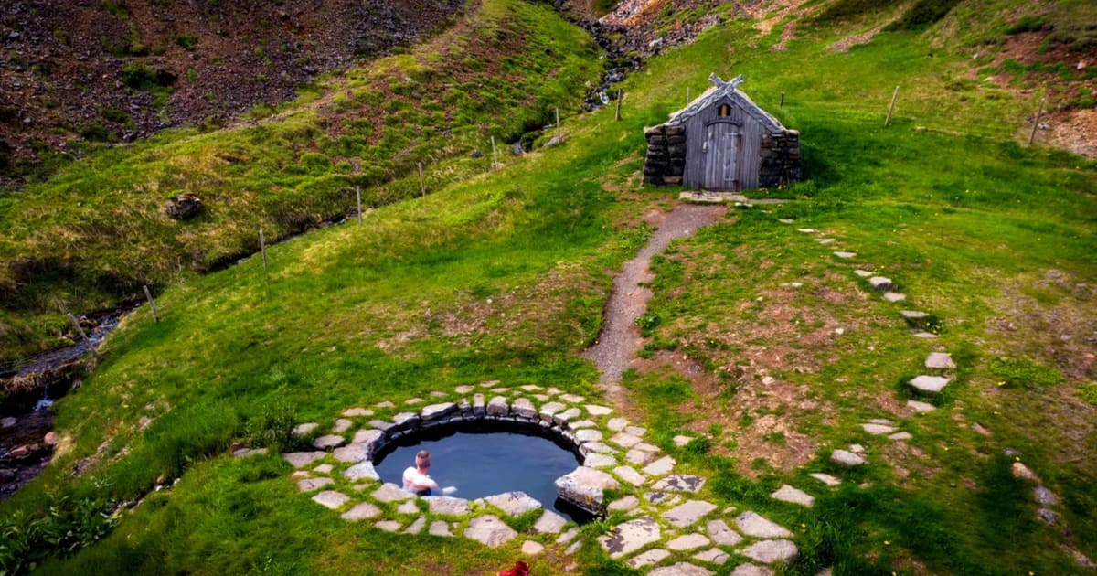 Guðrúnarlaug hot spring in Iceland with a round stone pool, lone bather, turf-roof hut and green valley framed by rocky hills
