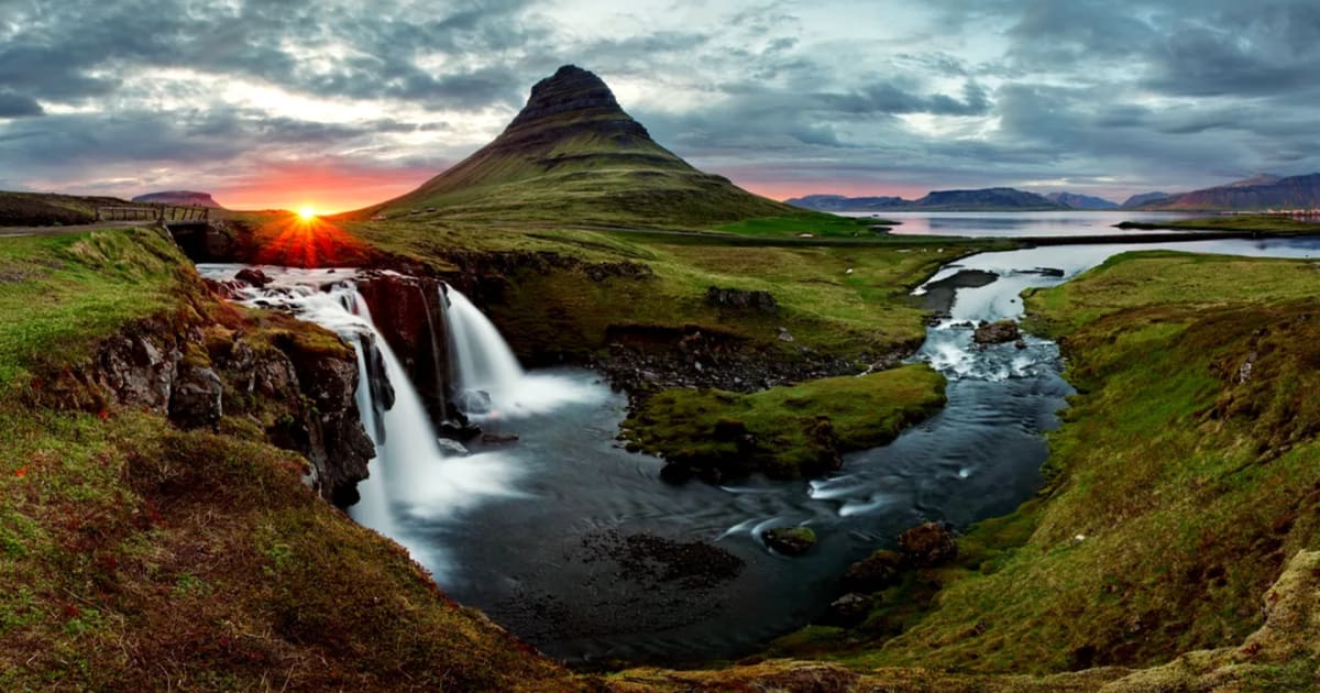 Midnight sun lights Kirkjufell and Kirkjufellsfoss waterfall in Iceland, with green June hills, river, clouds, and fjord views.