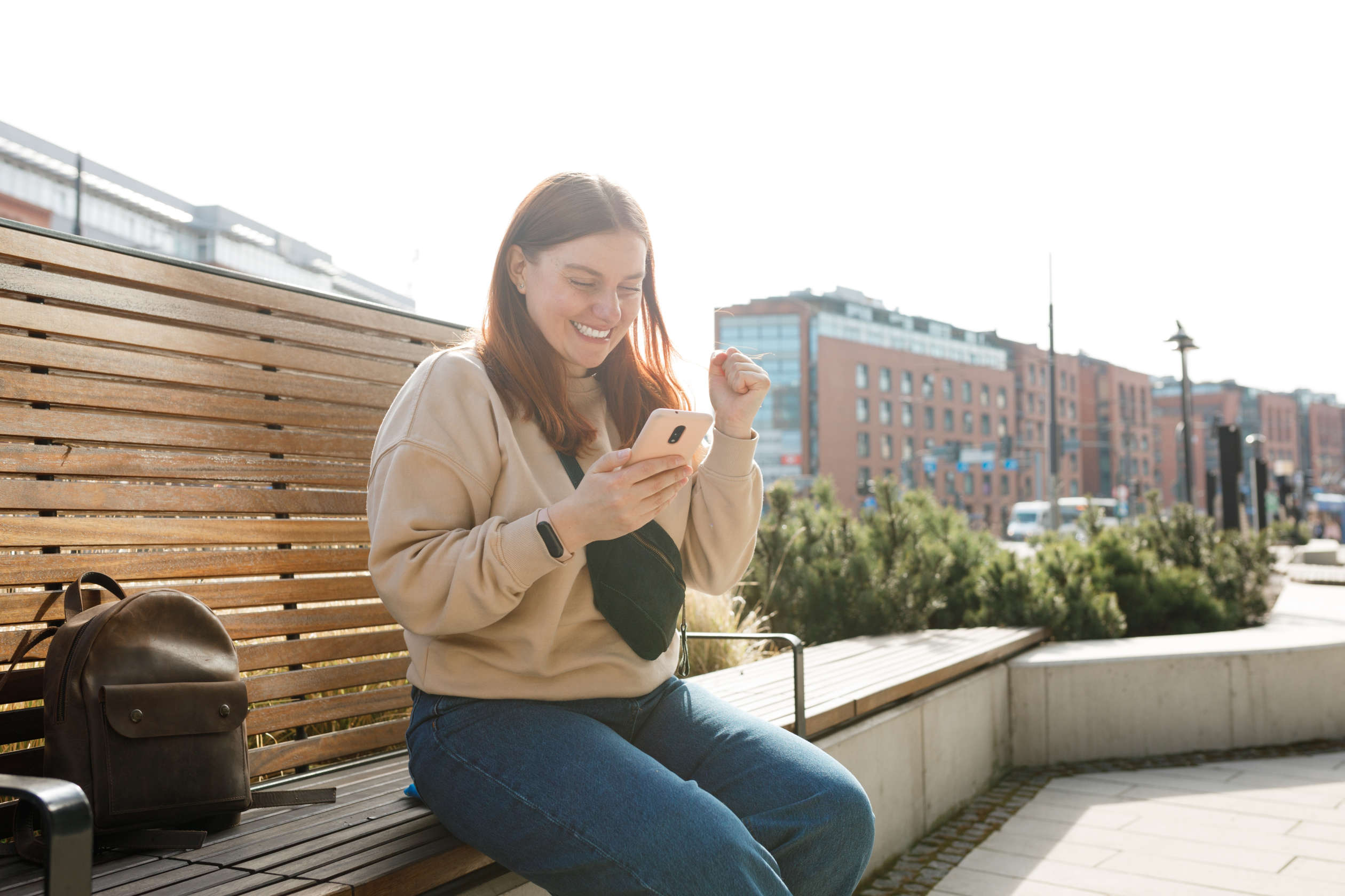 Woman checking her phone outdoors near a wooden fence, with city buildings in the background