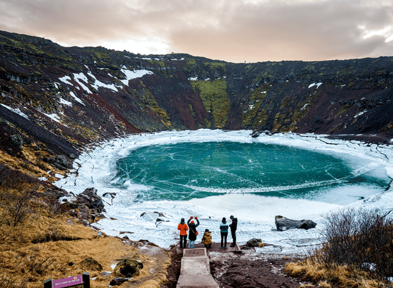 Visitatori che camminano lungo il bordo del cratere vulcanico di Kerið con un lago turchese sotto