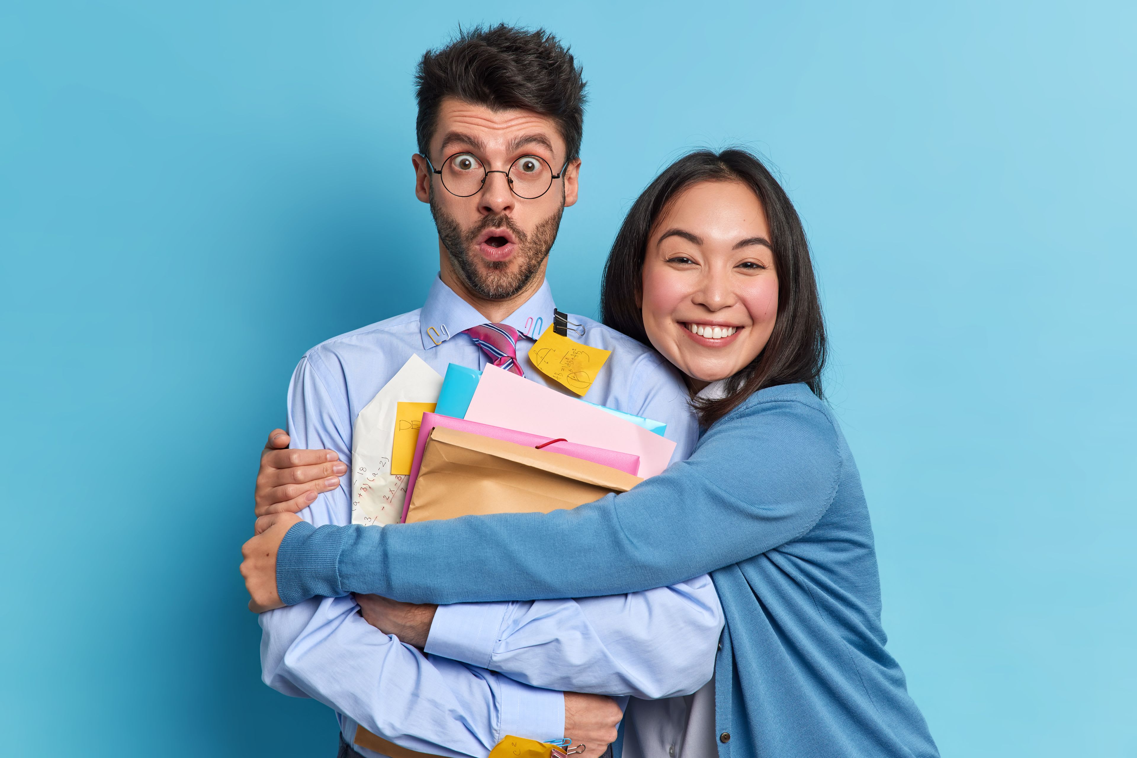 A colleague hugging another colleague who is holding files with a shocked look on his face