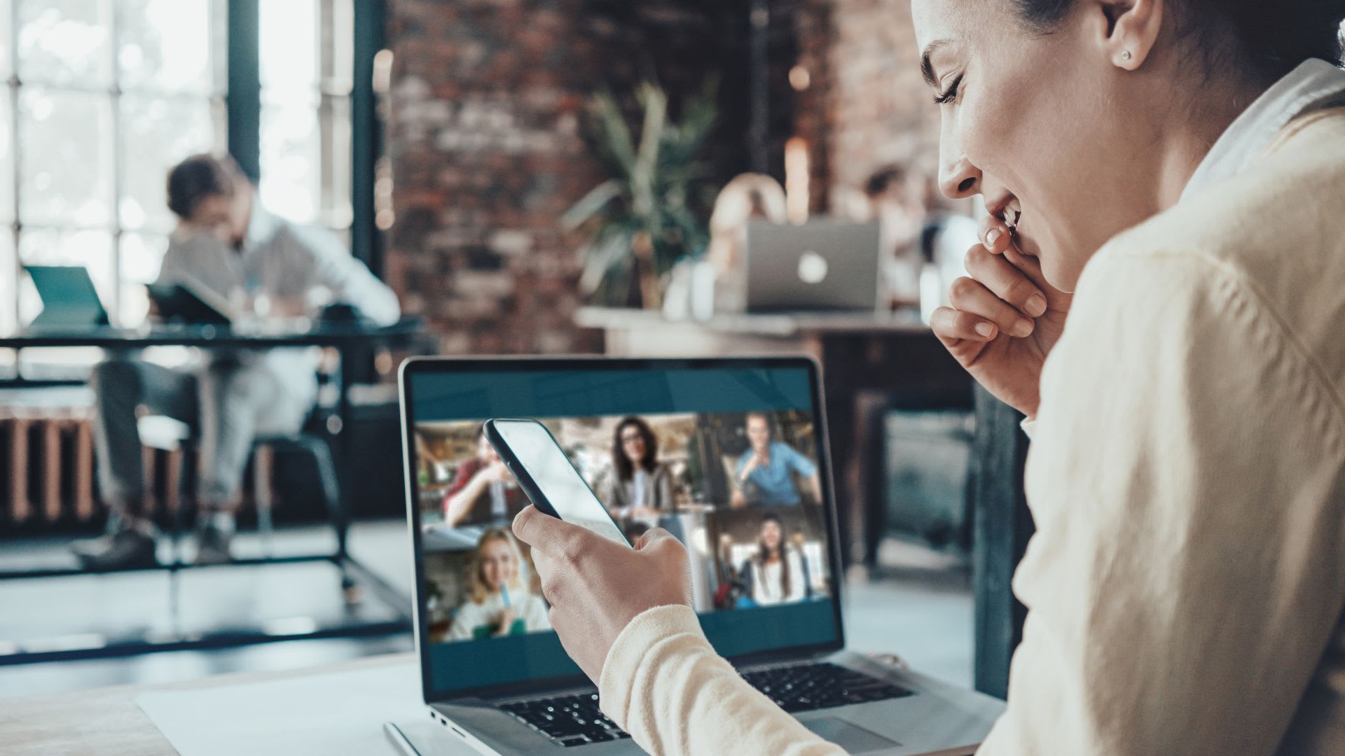 a person holding a phone and smiling while also on a remote meeting on their laptop
