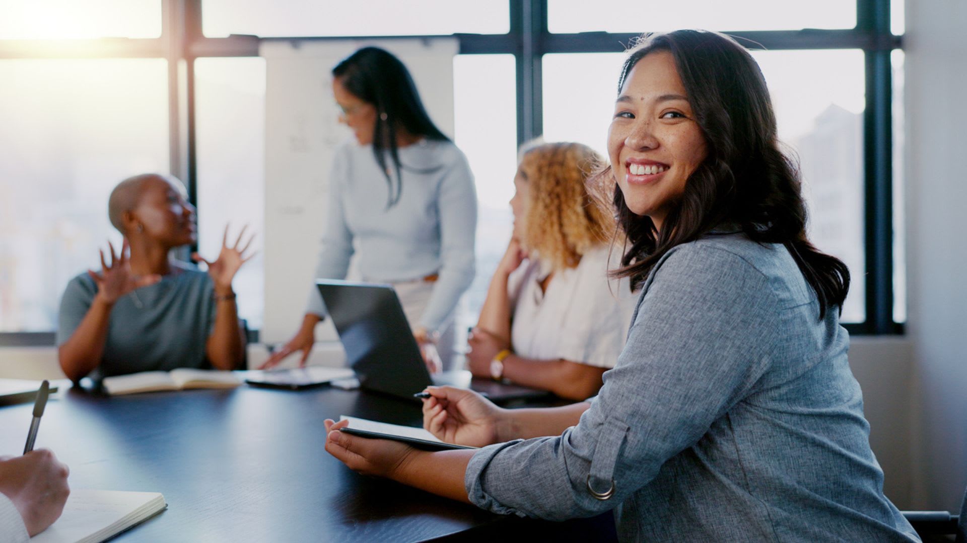 A group of work professionals sitting at a table, woman looking into camera smiling.