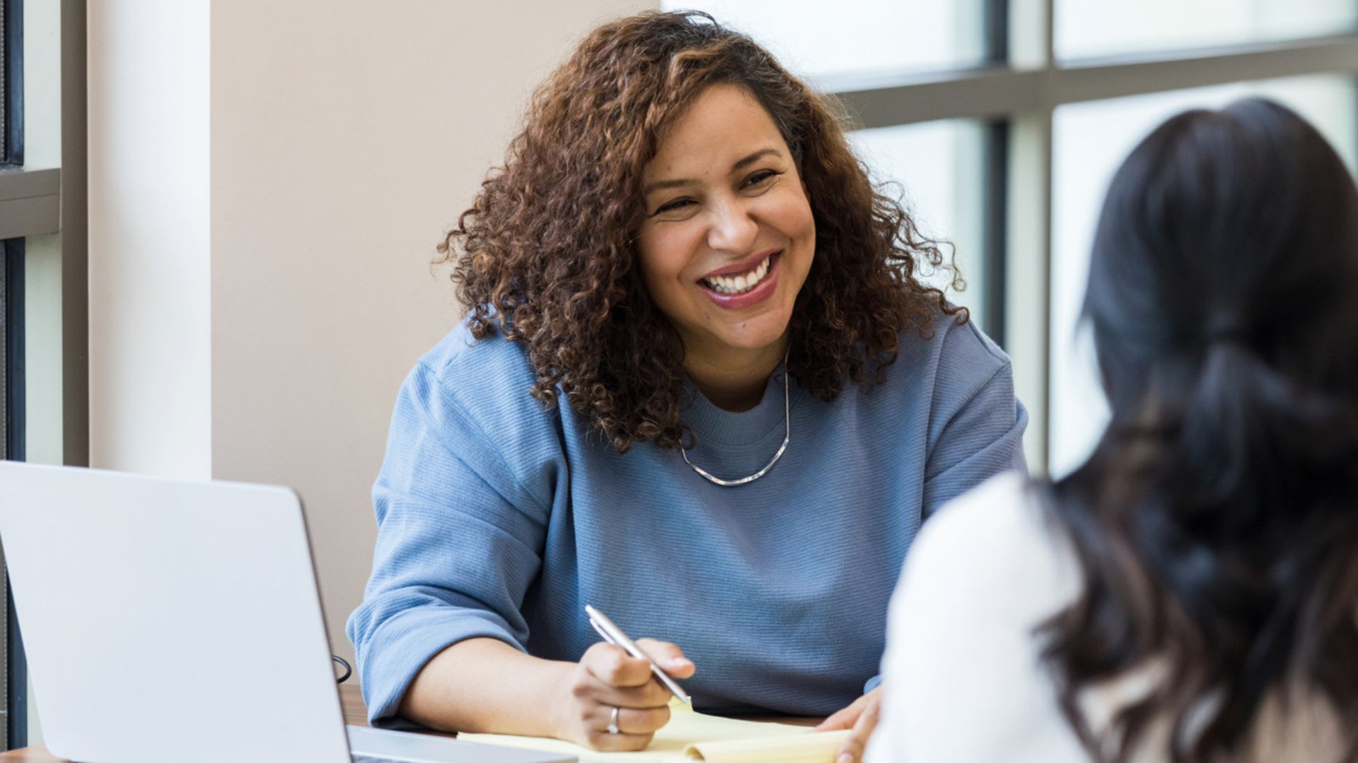 Smiling woman taking notes while engaged in conversation with another woman, representing a team manager engaging in a one on one meeting. 
