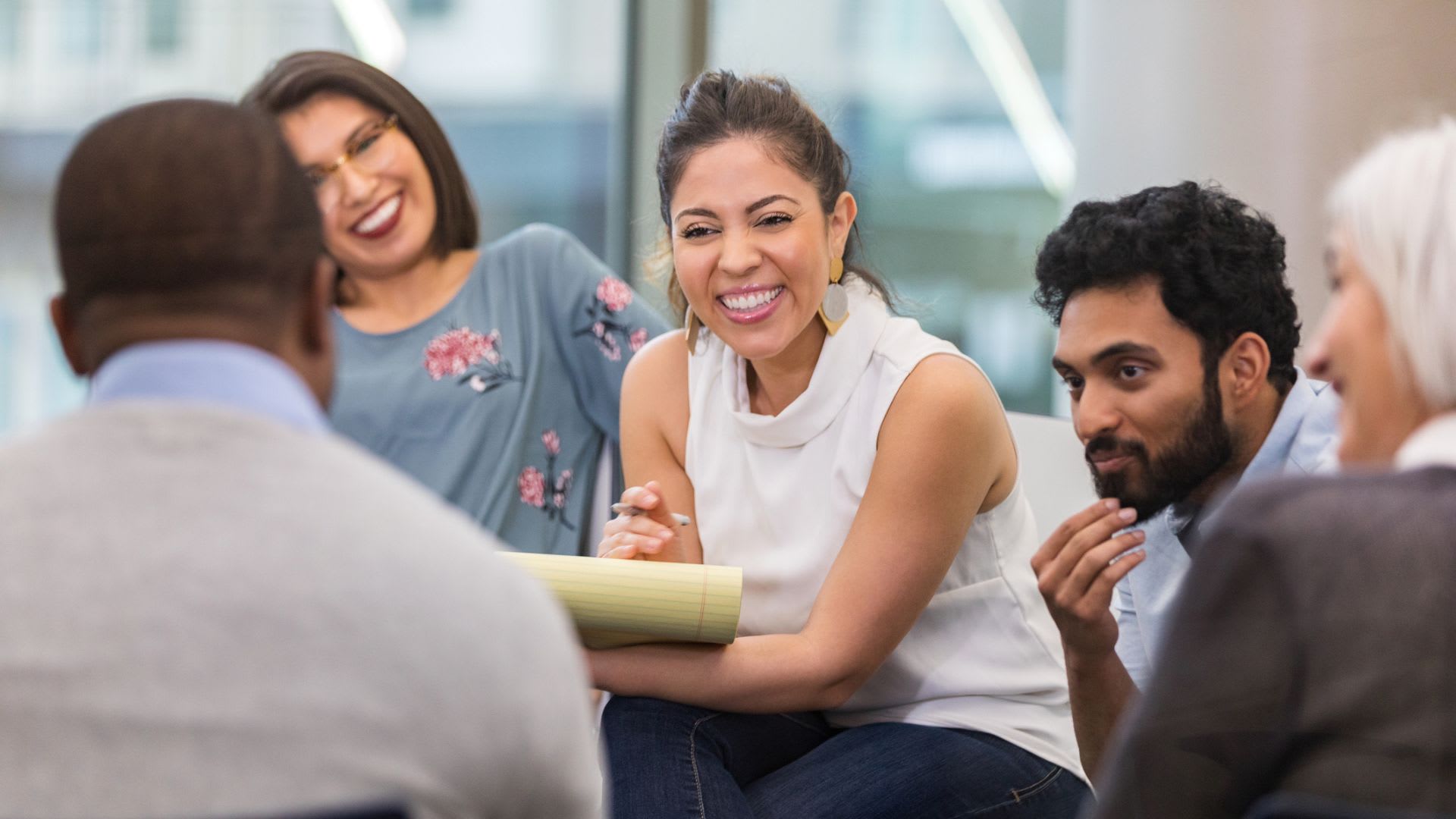 A diverse group of individuals engaged in conversation around a table, woman at center is smiling and intently listening to another team member