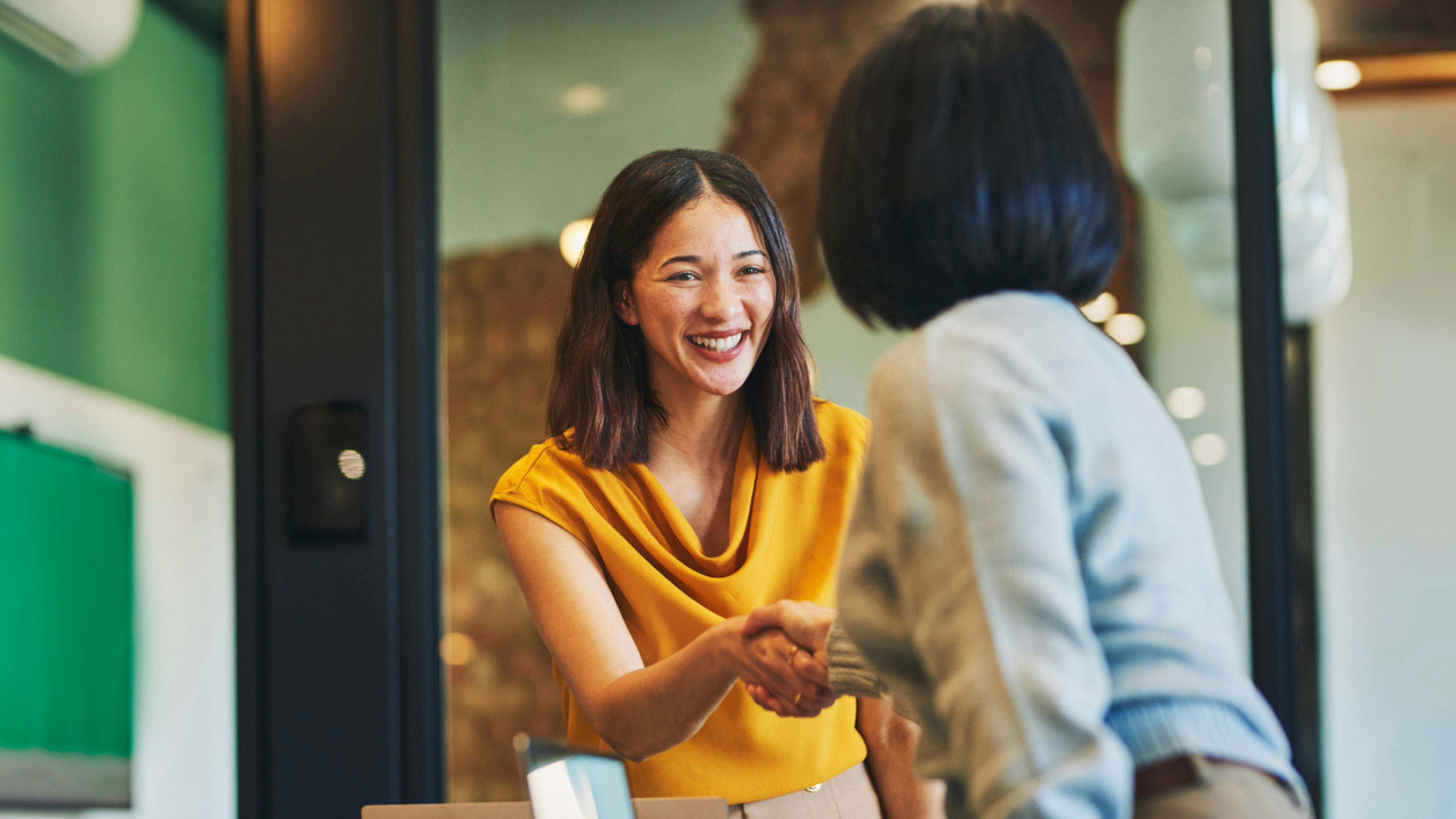 A woman and another woman shake hands in a professional office setting, symbolizing collaboration and agreement.