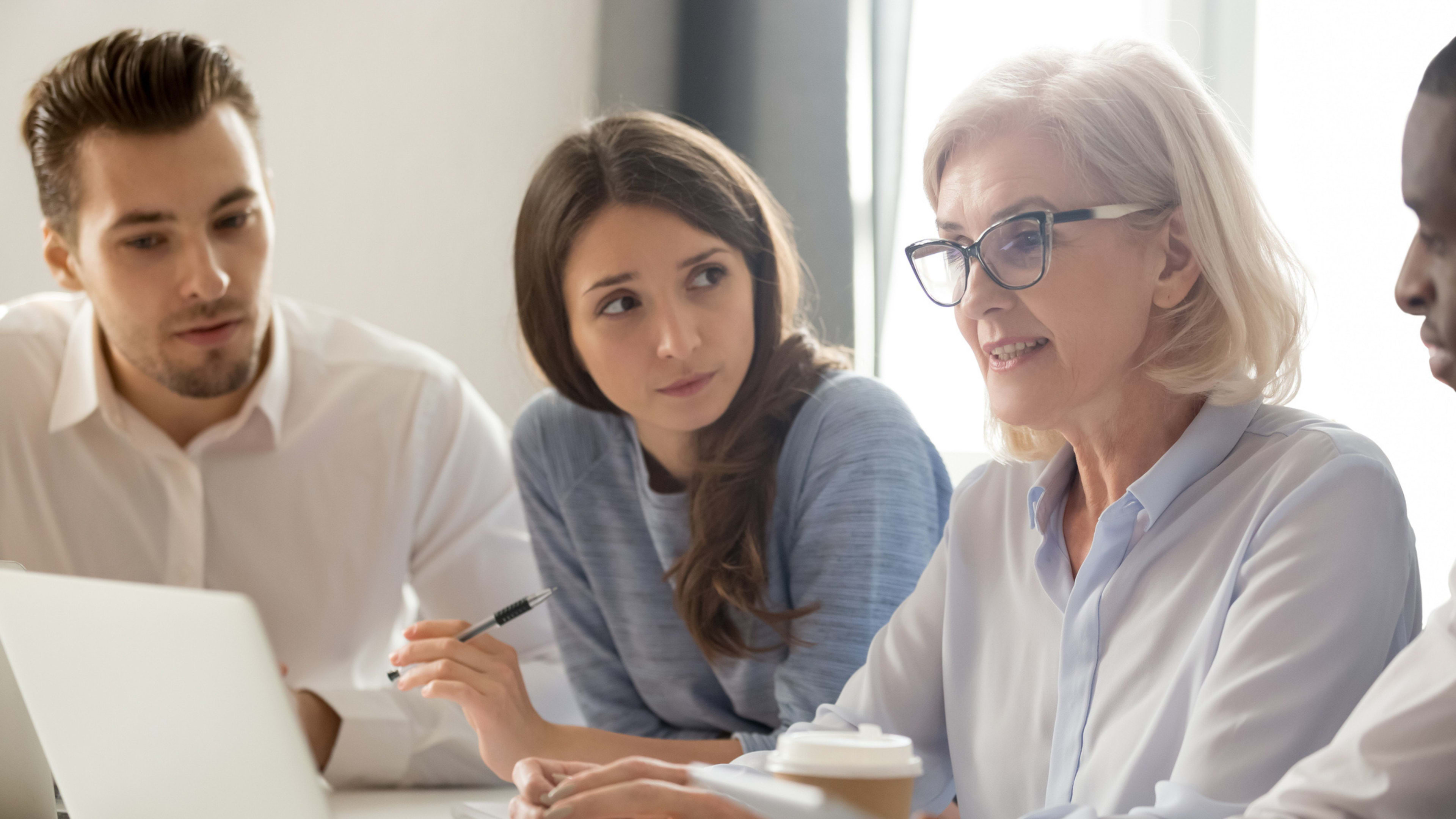 Three business professionals of different generations engaged in discussion at a table, demonstrating the current cross generational workplace. 