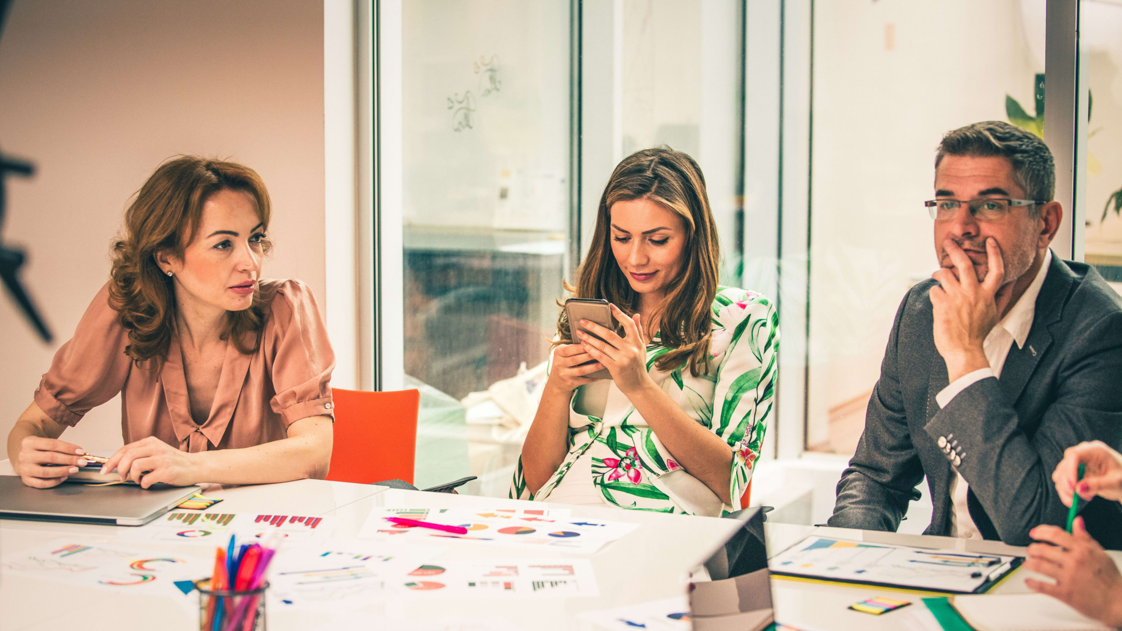 Three business professionals in a meeting at a conference table, they appear to be checked out and one is scrolling on her phone not paying attention. 