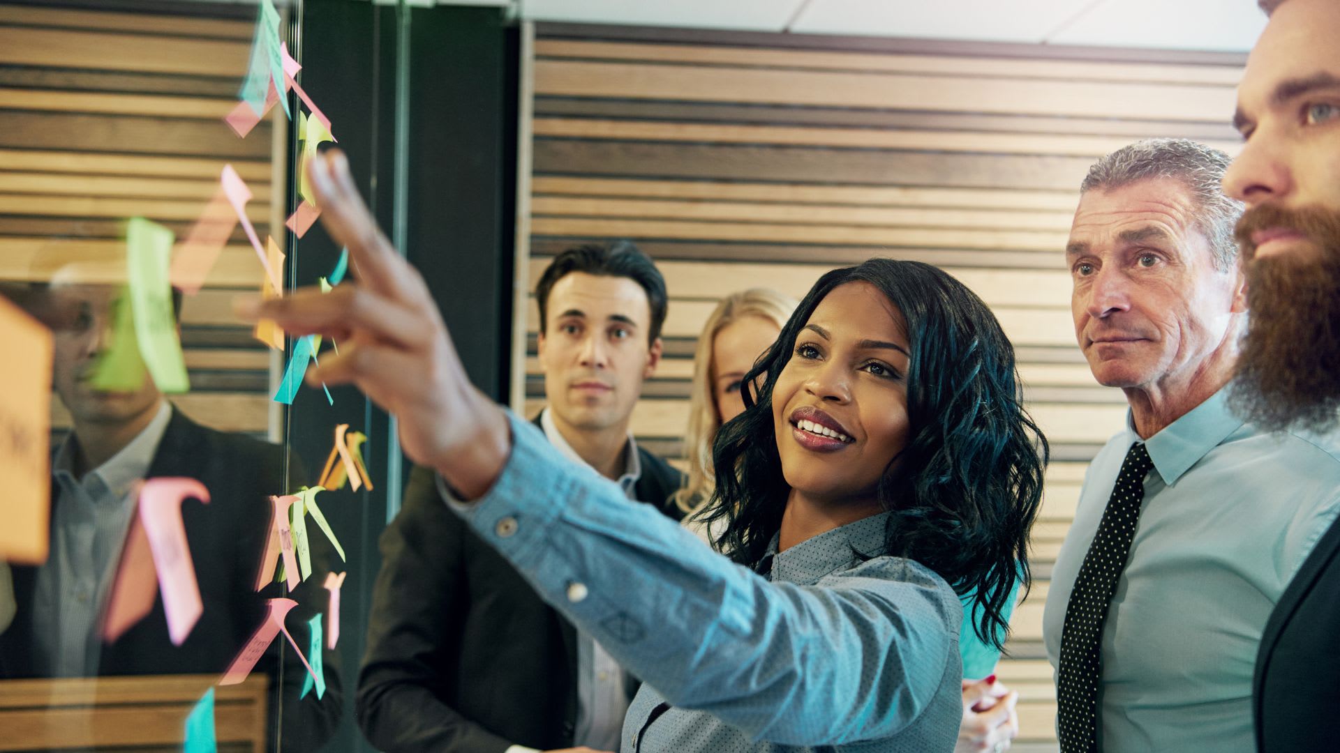 A diverse group of individuals examines a board filled with colorful sticky notes, engaged in discussion and collaboration.