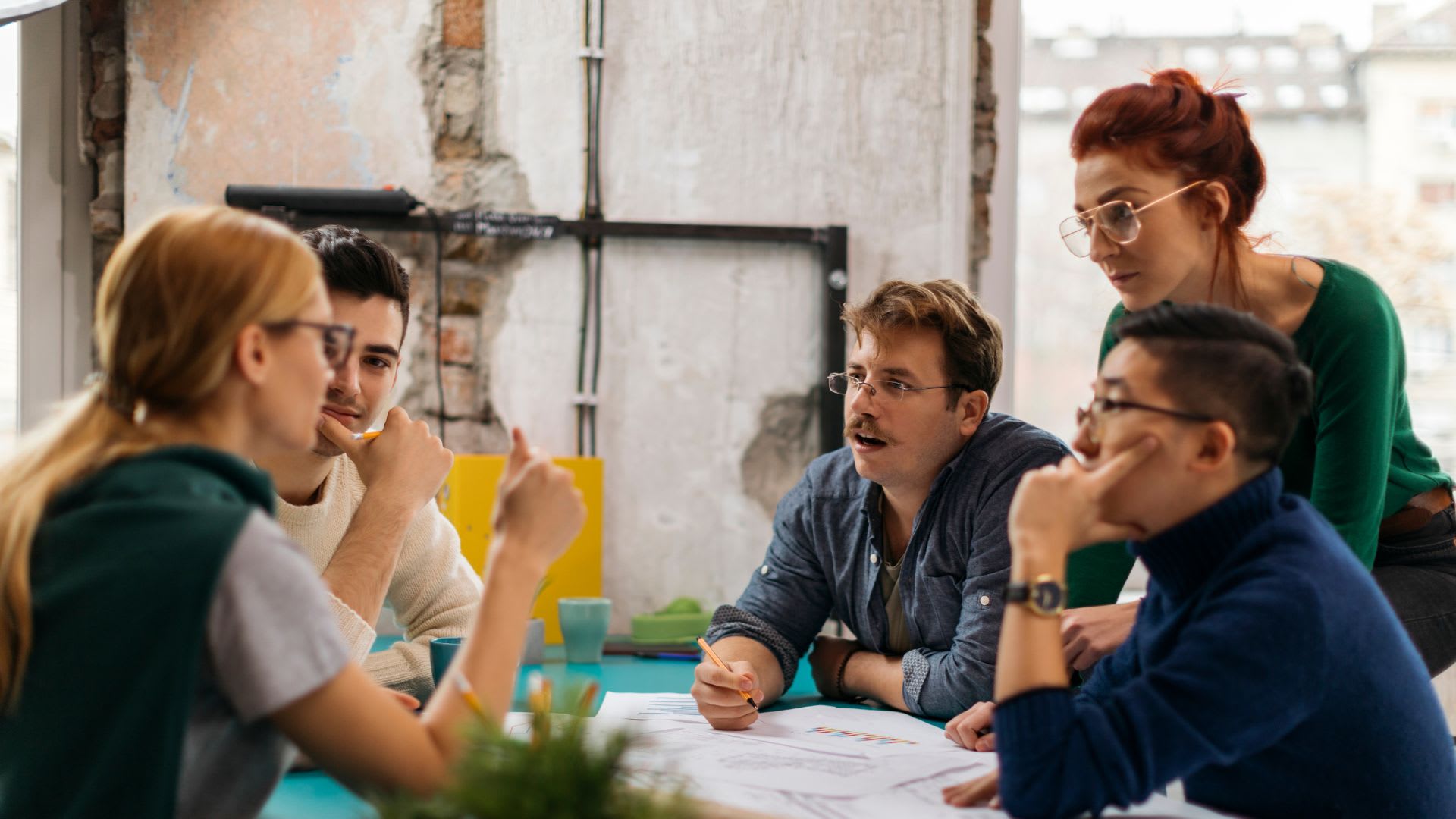 A diverse group of individuals collaborates around a table, engaging with a laptop in a productive meeting setting.