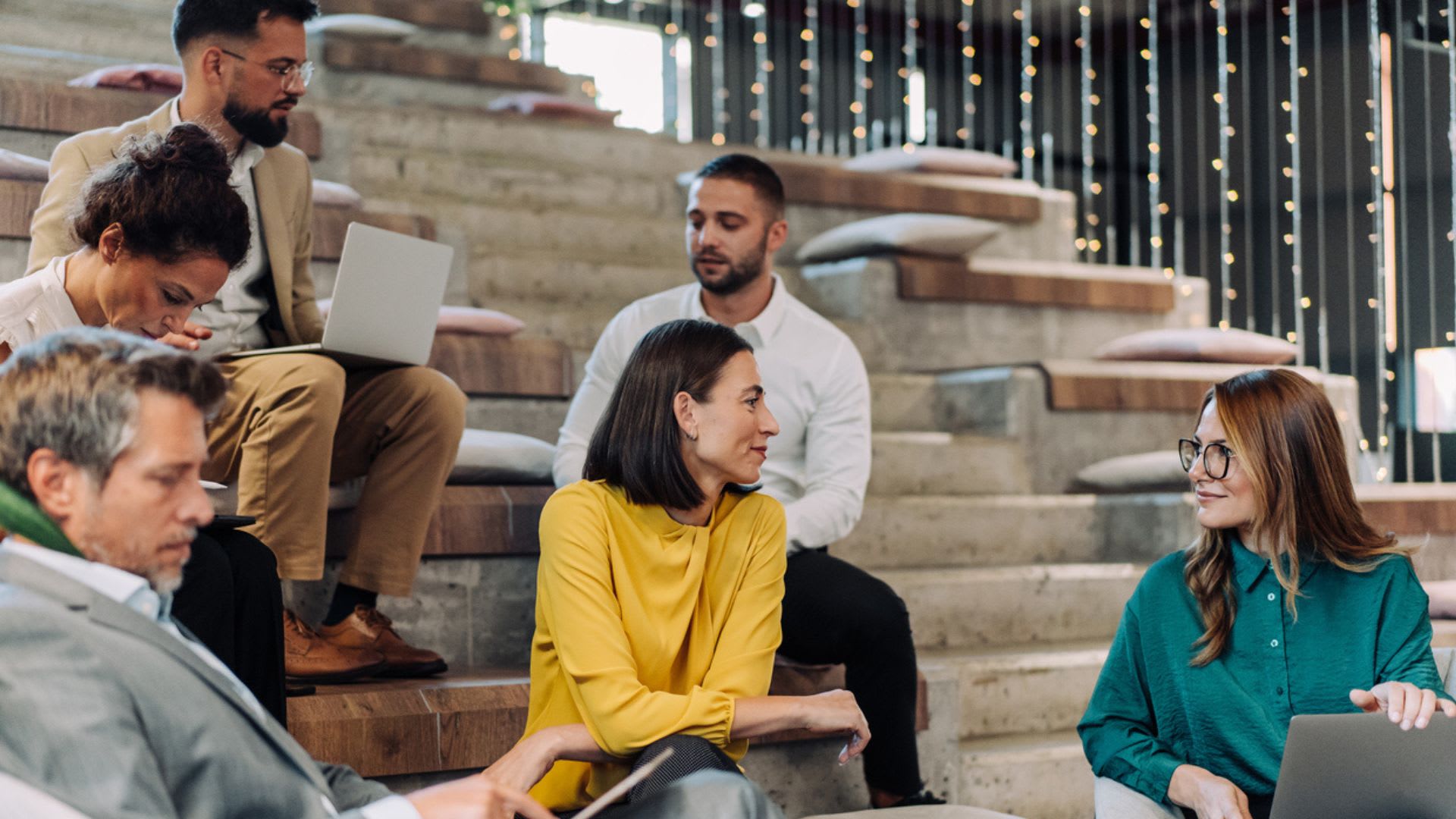 Shot of group of business people talking and using mobile devices together in modern office. Creative people with smart phones, tablets, laptops sitting together on stairs and having fun in their free time in the office. Business persons working together while sitting together on stairs in modern coworking space.