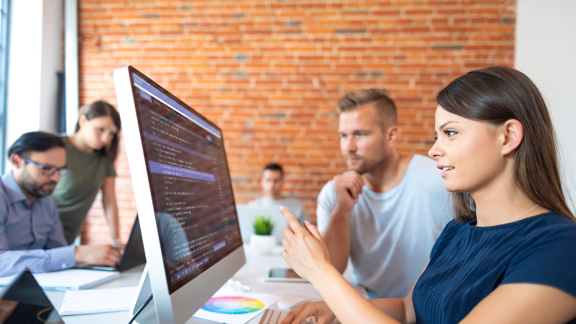 A woman sitting at a desk with a coworker, gesturing towards a computer screen, indicating something of interest or importance displayed on it.