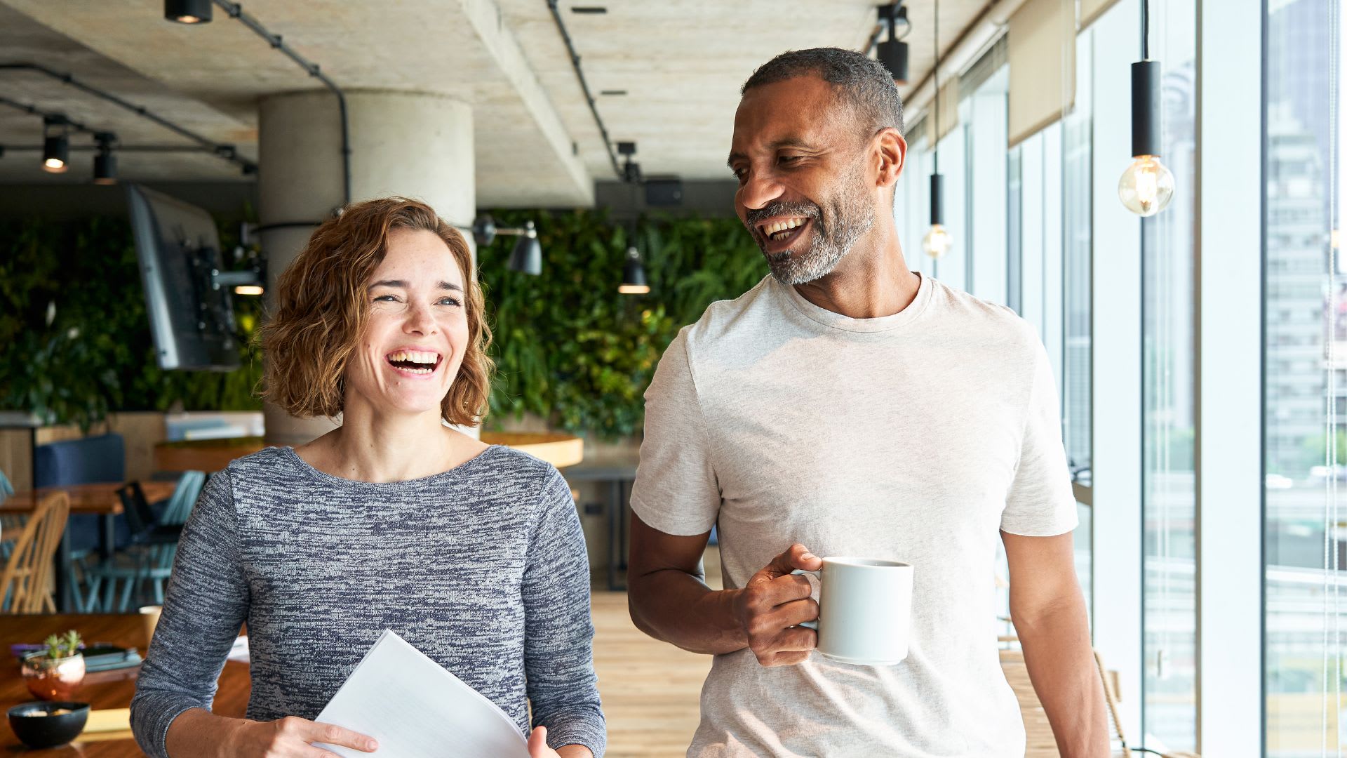 A man and woman stand in an office, each holding a coffee cup, engaged in conversation.