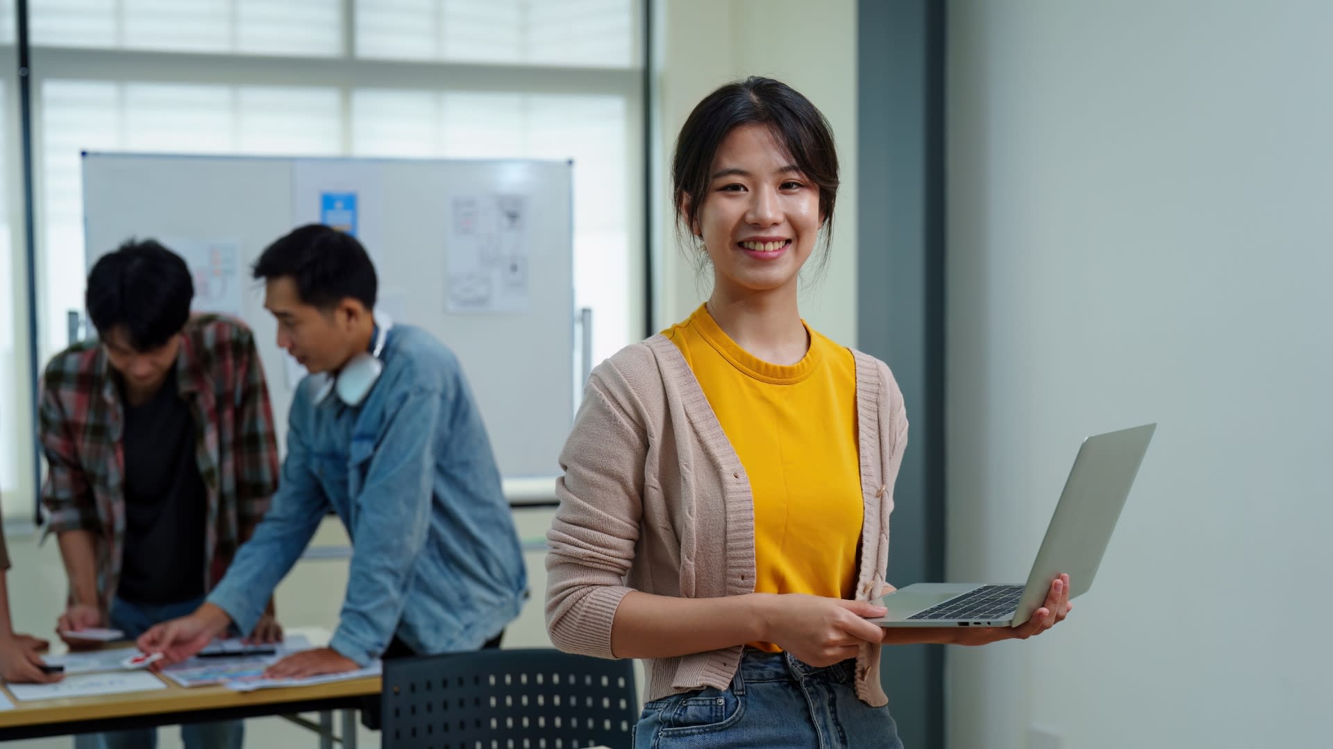 A woman holds a laptop in a workplace with coworkers working collaboratively behind her, she is looking at the camera and smiling. 