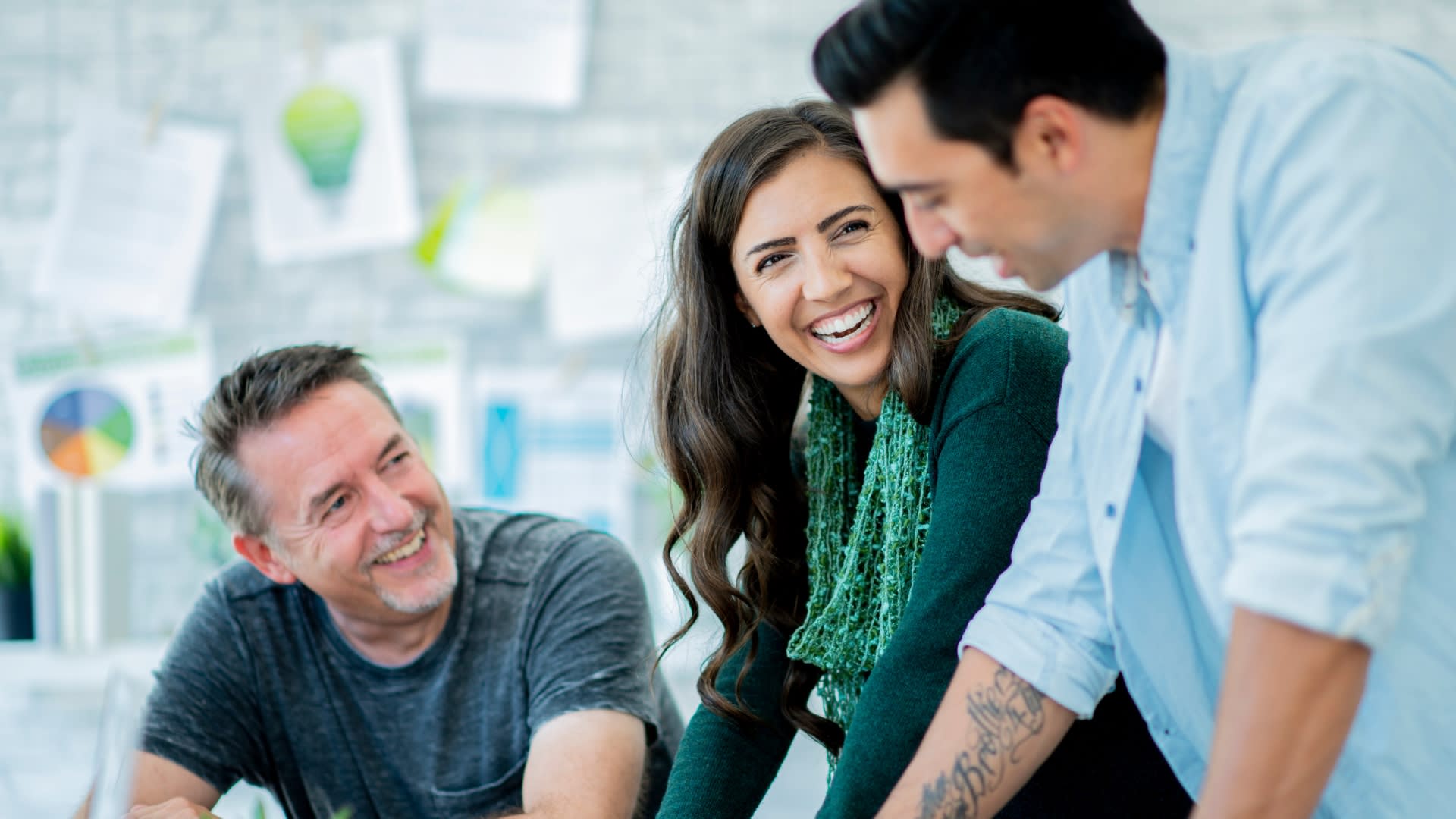 Three smiling individuals gathered around a table, with a laptop placed in the center, demonstrating a sense of camaraderie.