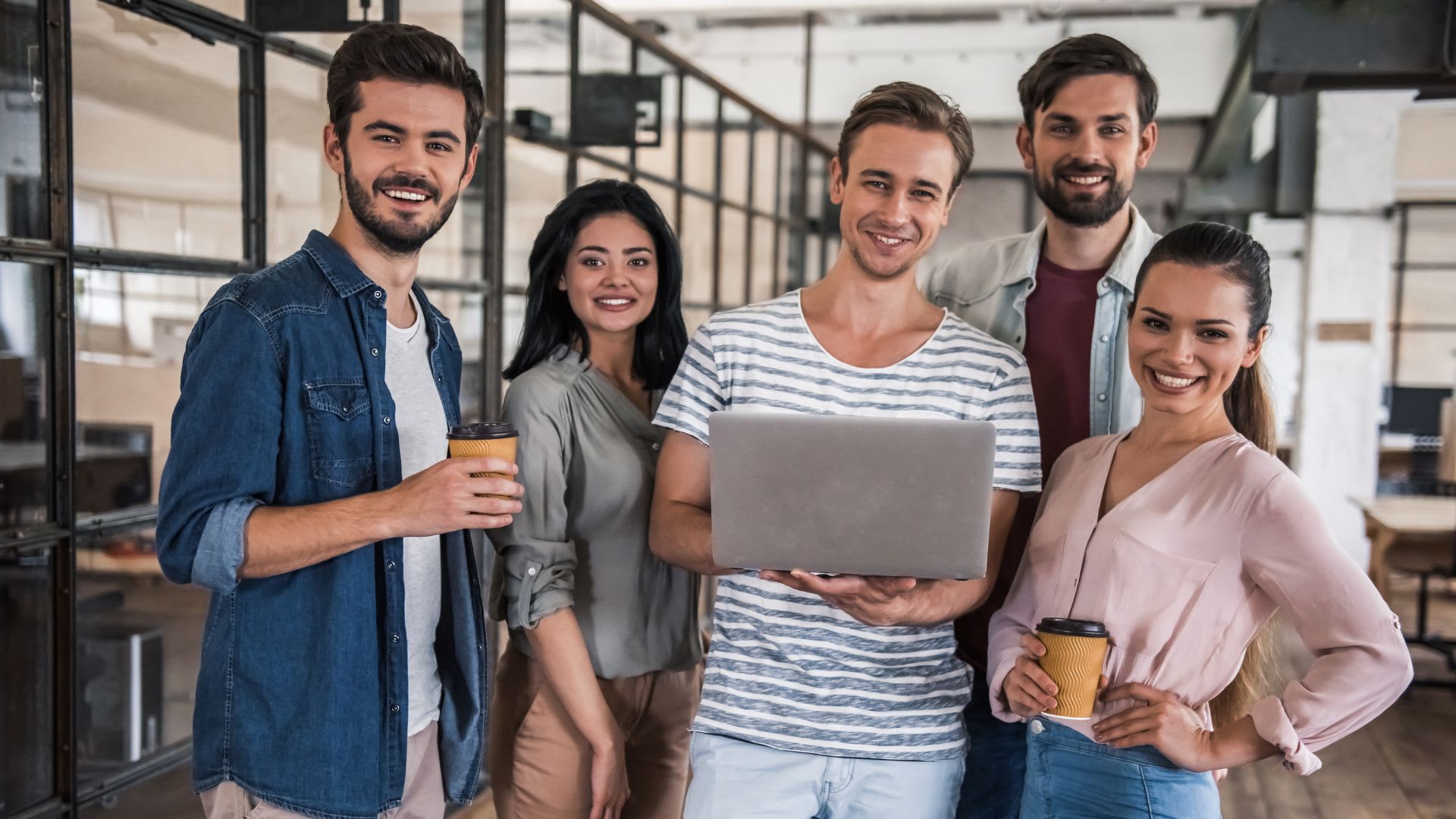 A group of professional coworkers in a modern work environment, standing together and looking at camera smiling. 
