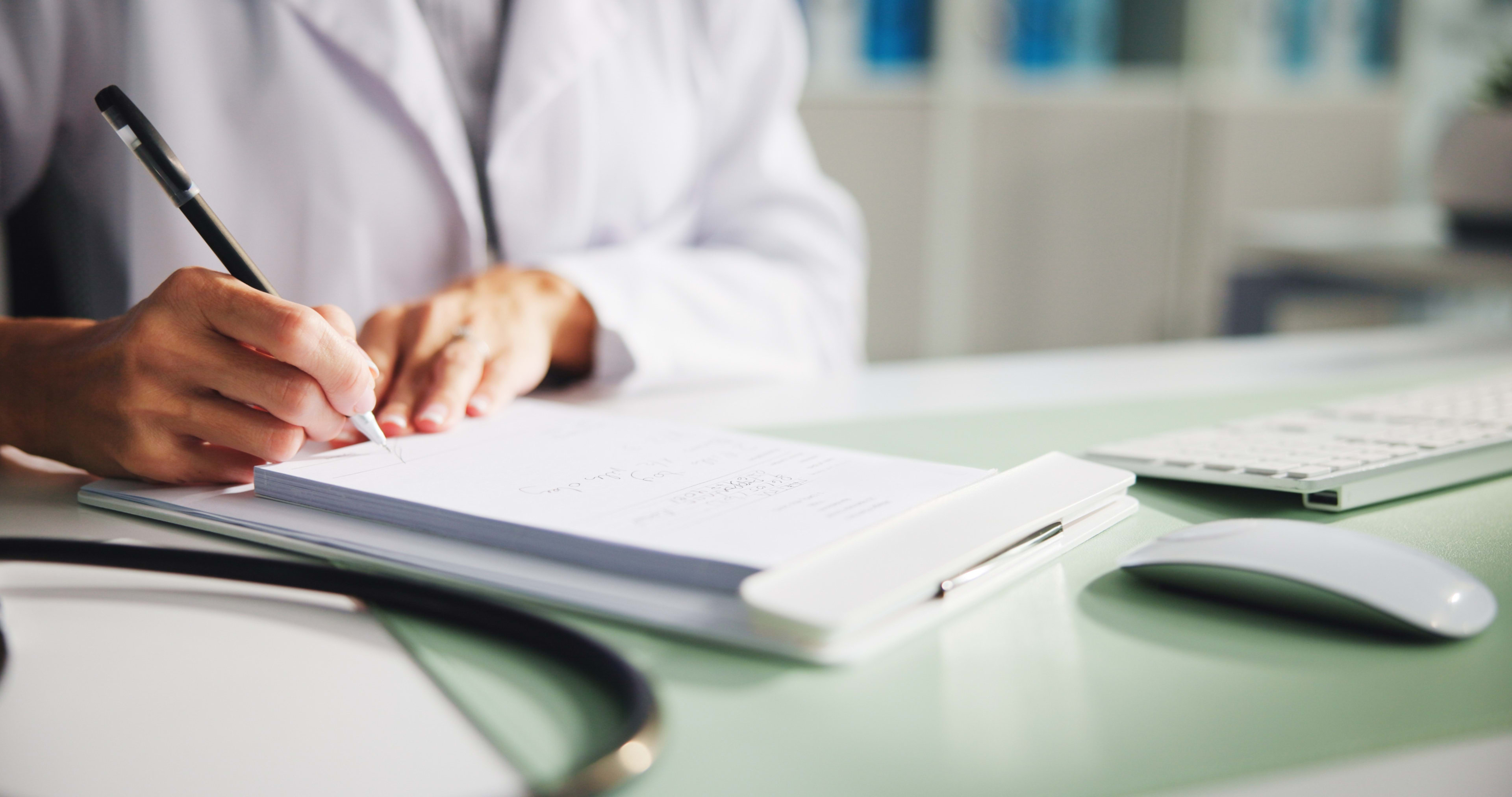 A person in a white coat writes on a clipboard at a desk with a stethoscope, keyboard, and mouse nearby.