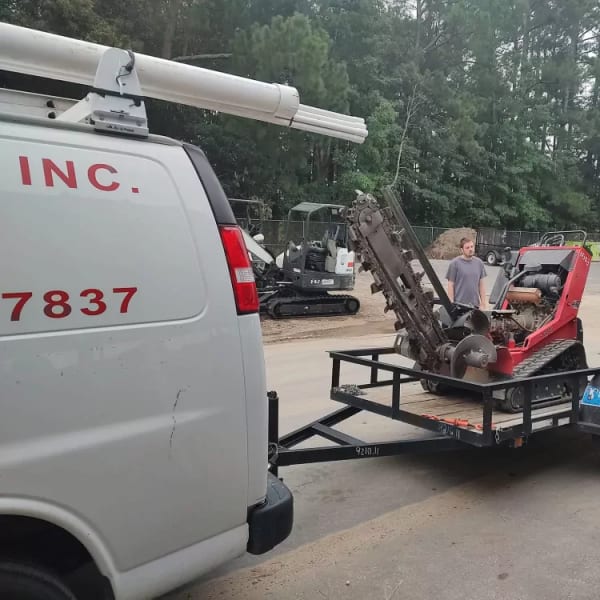 Rosewood Plumbing service van and crew loading a walk-behind trencher onto a trailer