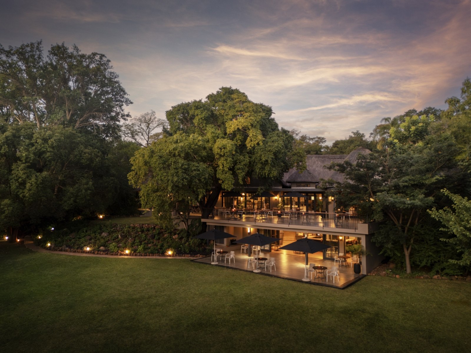 The hotel's restaurant terrace lit up at dusk, set among tall trees above a sweeping lawn