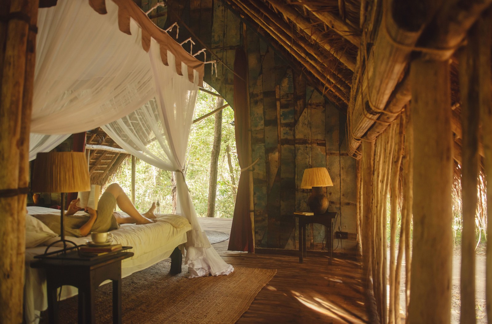 A person relaxes on a white canopy bed inside a rustic, thatched-roof wooden room overlooking a forest.