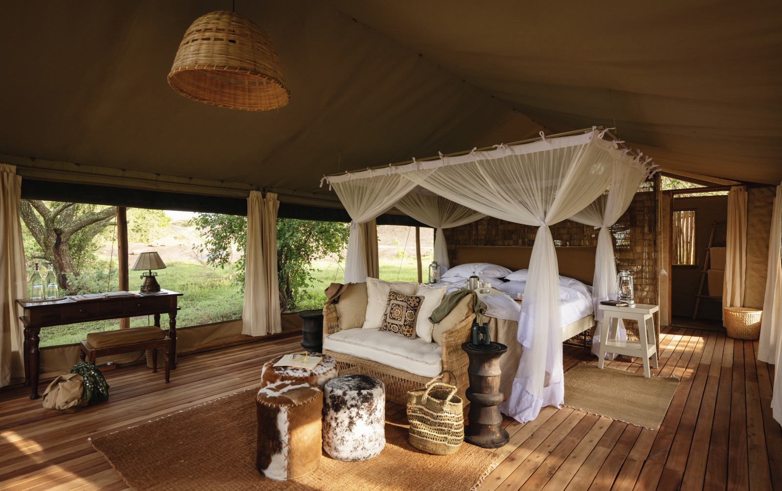 Interior of a luxury safari tent with a large canopy bed, mosquito netting, and wicker furniture overlooking a green savanna.