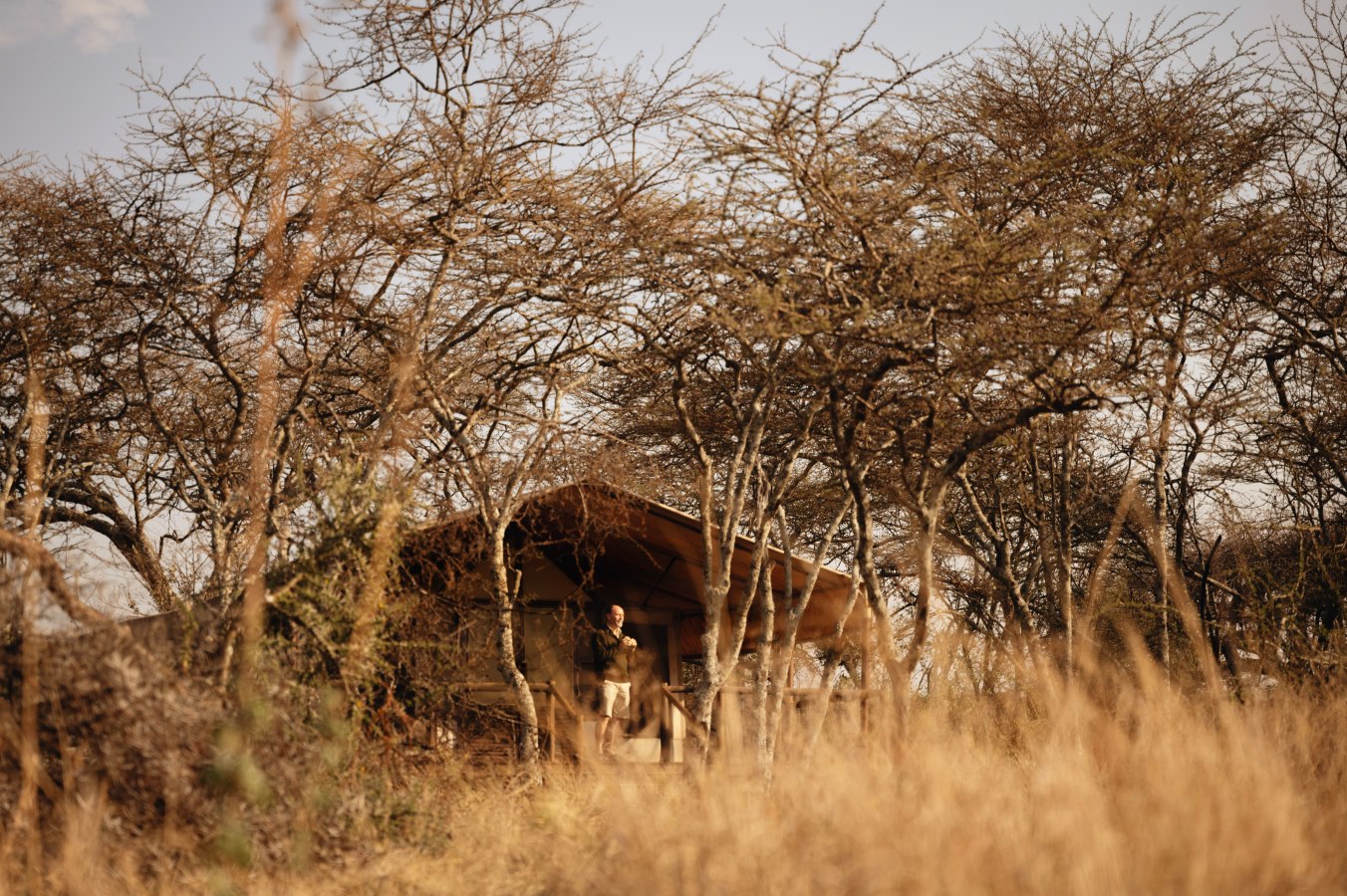 A man standing on the porch of a safari tent in a dry savanna landscape.