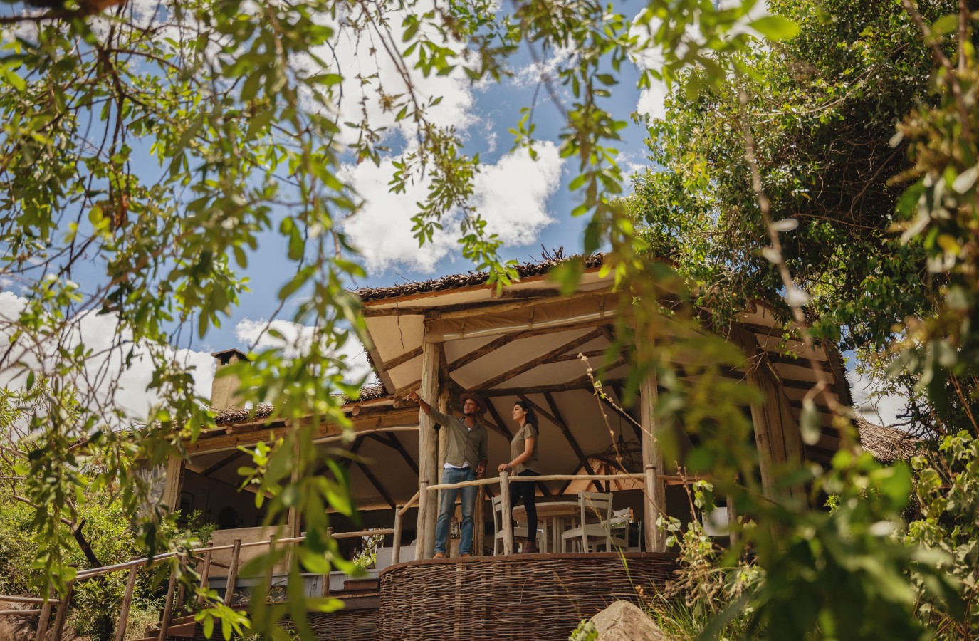 A man and woman stand on the wooden deck of a safari lodge, framed by leafy green branches, looking out at the surrounding nature.