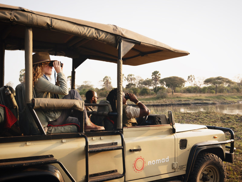 A group of people on a safari tour in a beige open-top vehicle, looking out over a river at golden hour.