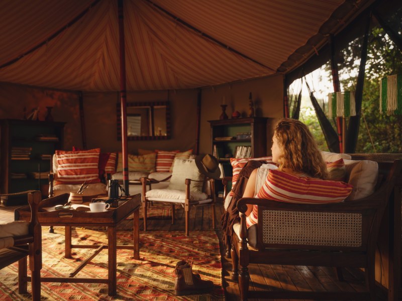 Interior of a luxurious safari tent at sunset featuring a woman relaxing in a wooden chair among warm-toned furnishings and striped pillows.