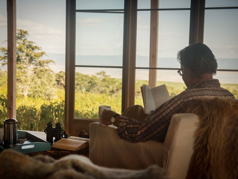 A man sits in a cozy armchair reading a book by a large window overlooking a vast green savanna.