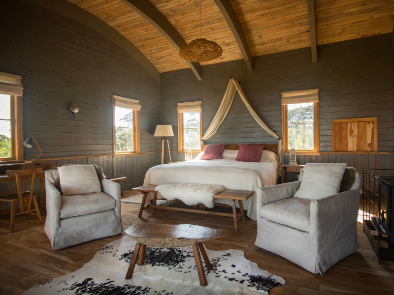 A luxurious rustic bedroom featuring dark wood-paneled walls, a canopy bed with pink pillows, and comfortable white armchairs on a cowhide rug.