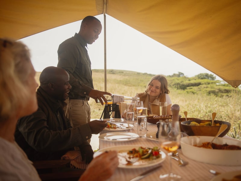 A waiter serves guests dining under a large canvas tent in an open savanna landscape.
