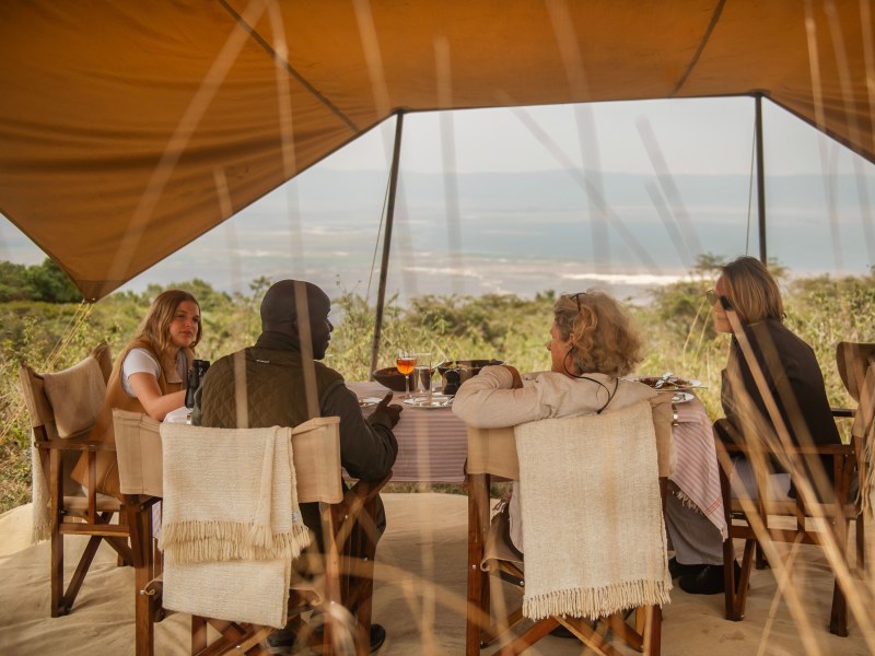 A group of four people enjoying a meal under a large tan safari tent with an expansive view of the African plains.