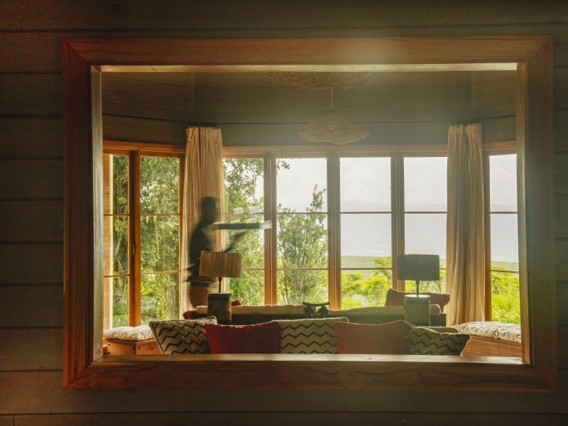 A warm, interior view of a luxury lodge lounge area framed by a wooden border, featuring patterned cushions and large windows overlooking a lush green valley.