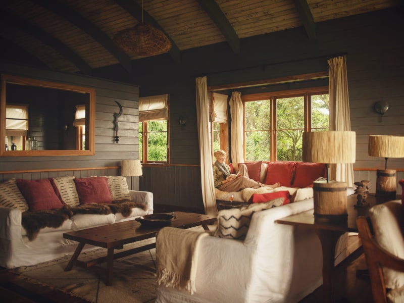 A cozy, rustic living room with dark wood walls, white slipcovered sofas, and a woman reading on a sunlit window seat.