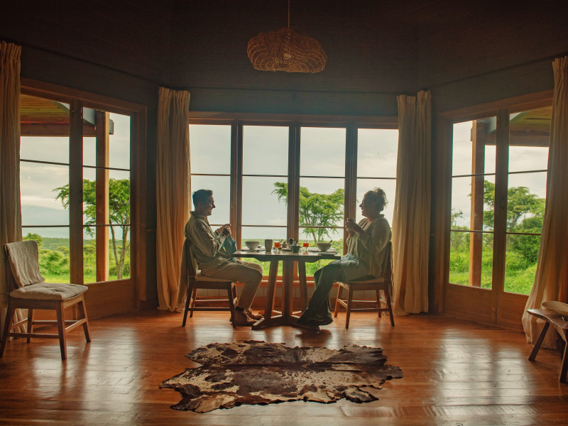 A man and a woman sitting at a dining table inside a room with large windows looking out onto a green savanna.
