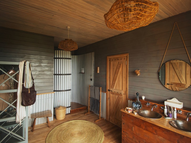 A rustic-style bathroom with wooden walls, corrugated metal accents, a wooden double vanity with metal sinks, and a round mirror.