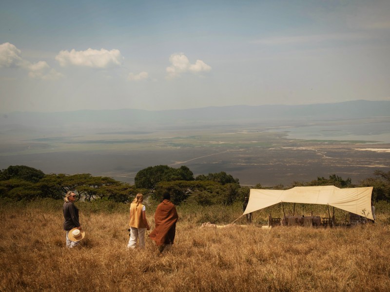 Three people walking across a dry grassy field towards a white canopy tent overlooking a vast valley and lake.