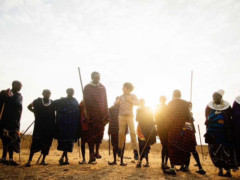 A young boy jumps high in the air while surrounded by Maasai people in traditional red and blue shukas during sunset on the savanna.