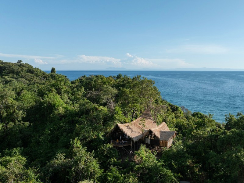 An eco-lodge with traditional thatched roofs nestled in a dense green forest on a hillside overlooking a calm blue sea.
