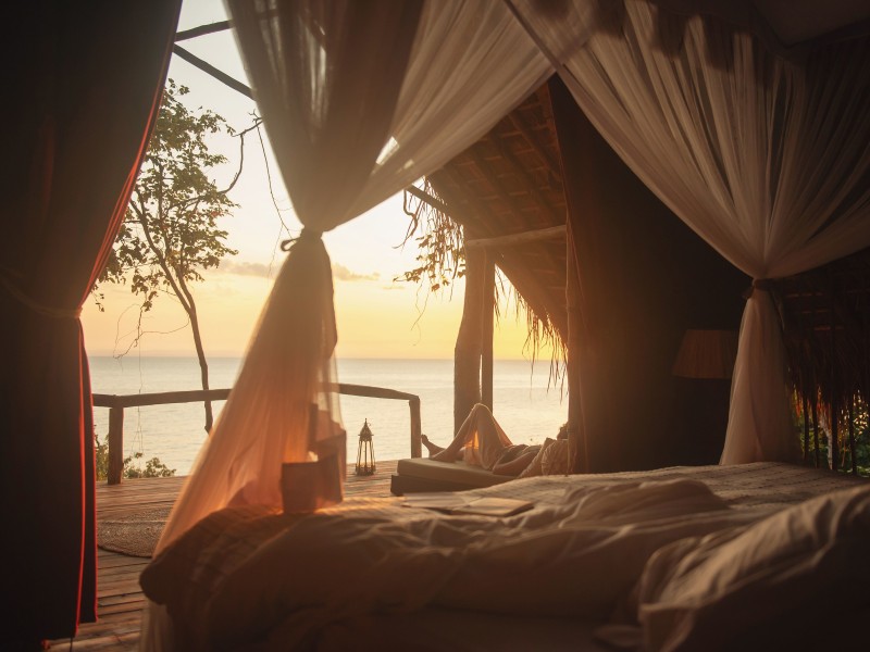 A serene view from a rustic bedroom looking out onto a wooden deck and the ocean during sunset.