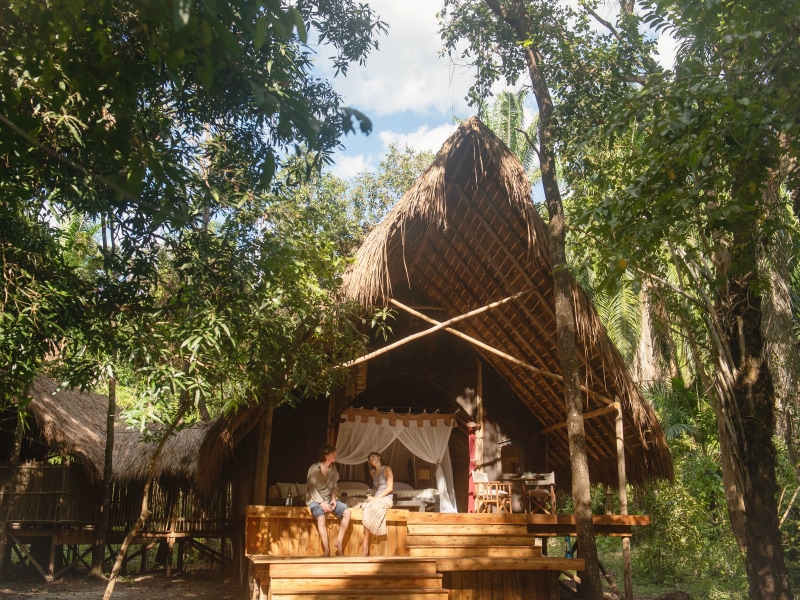 A couple sits on the wooden steps of a luxury thatched-roof lodge nestled in a lush jungle.